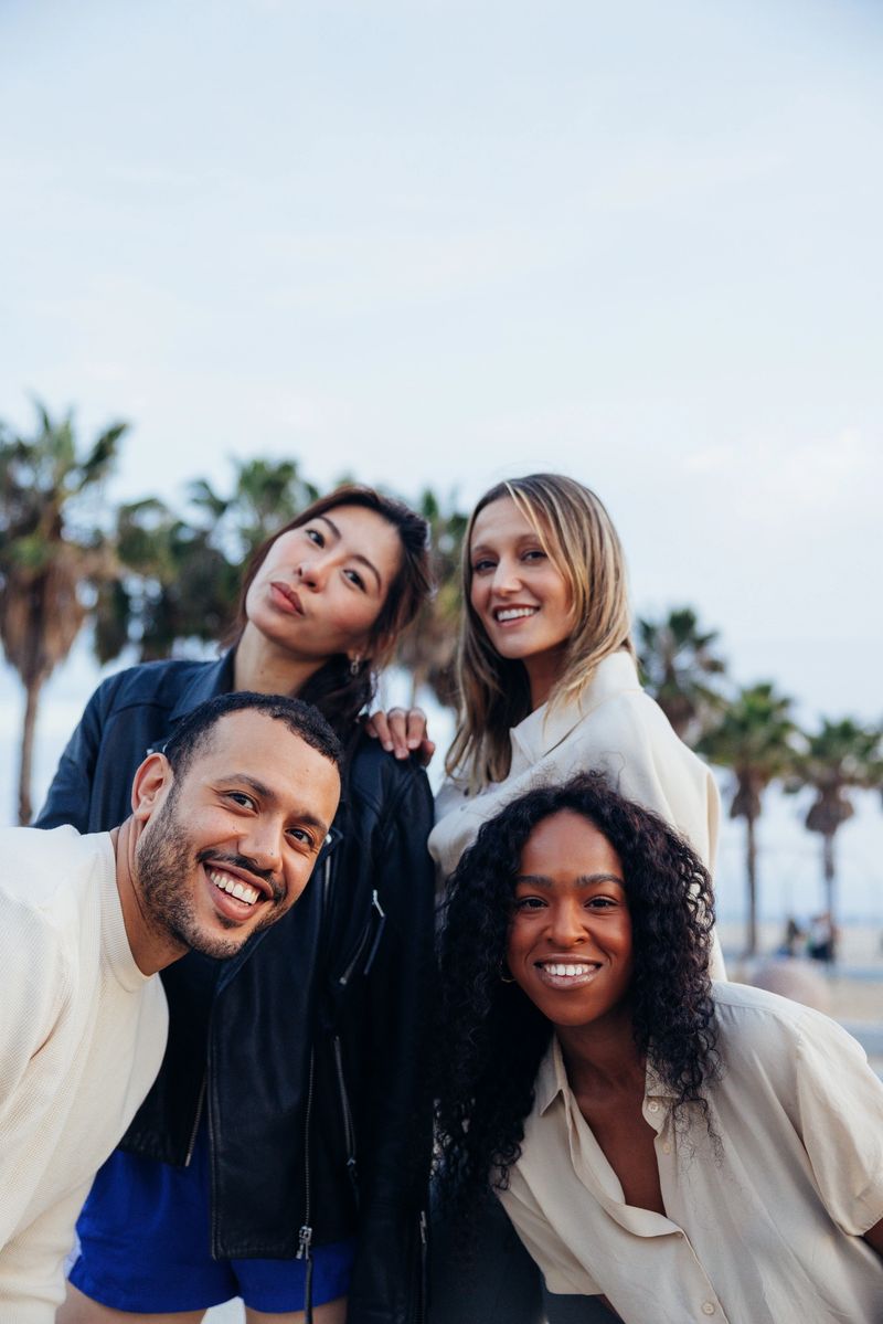 A diverse group of four young adults standing close together outdoors in a modern city environment. They face the camera with relaxed, natural body language, suggesting familiarity and social connection rather than a staged pose. Daylight illuminates the scene evenly, while the urban backdrop provides contemporary context for a lifestyle portrait centered on friendship, inclusion, and shared presence.