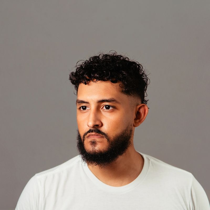 Hispanic man with curly hair and beard looking away, considering male grooming, beauty, and diet for healthy skin