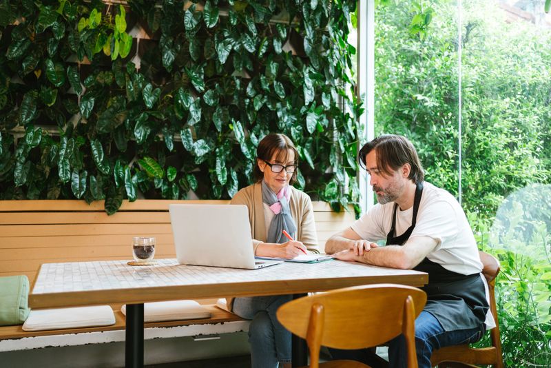 Two people sit at a table with a laptop, discussing work in a bright, green indoor setting with plants on the wall.
