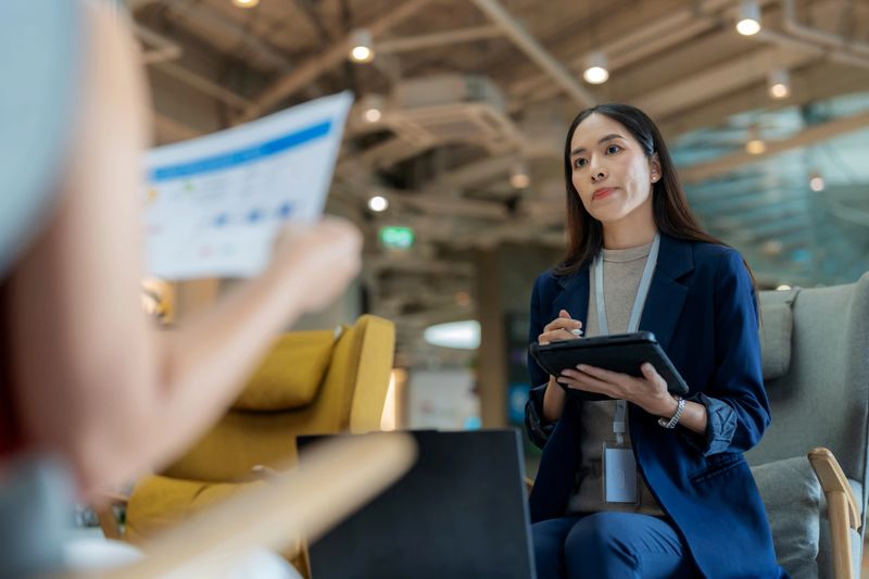 Professional Asian female marketers analyzing sales graphs and financial reports in a modern office. Young businesswomen discussing marketing strategy and taking notes on a tablet during a productive teamwork meeting.