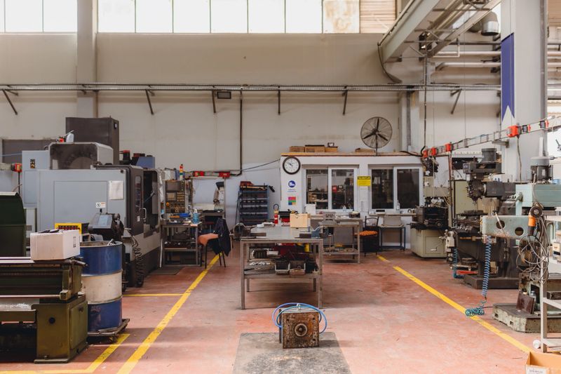 A wide interior view of an industrial manufacturing workshop showing CNC machines, milling equipment, workbenches, tools, and metalworking machinery arranged in an organized production environment, representing precision engineering, factory operations, mechanical industry, and modern manufacturing processes with copy space
