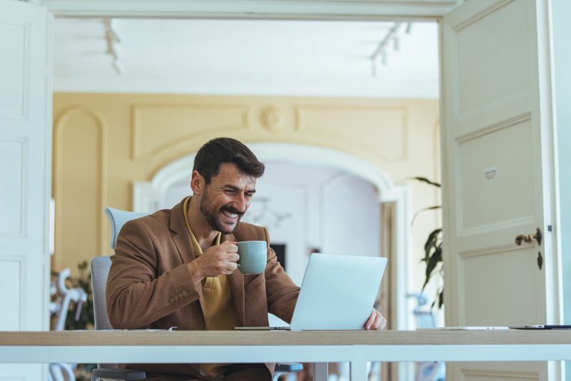 A cheerful man in a brown blazer sits at a sunlit office desk, using a laptop and sipping coffee, conveying a productive, casual business mood in a modern workspace.
