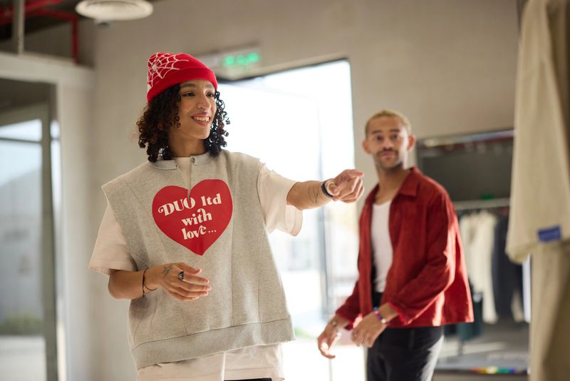 Smiling woman pointing at dress while shopping in modern clothing store. Customer is browsing fashion items with male friend. She is wearing stylish casuals along with knit hat.