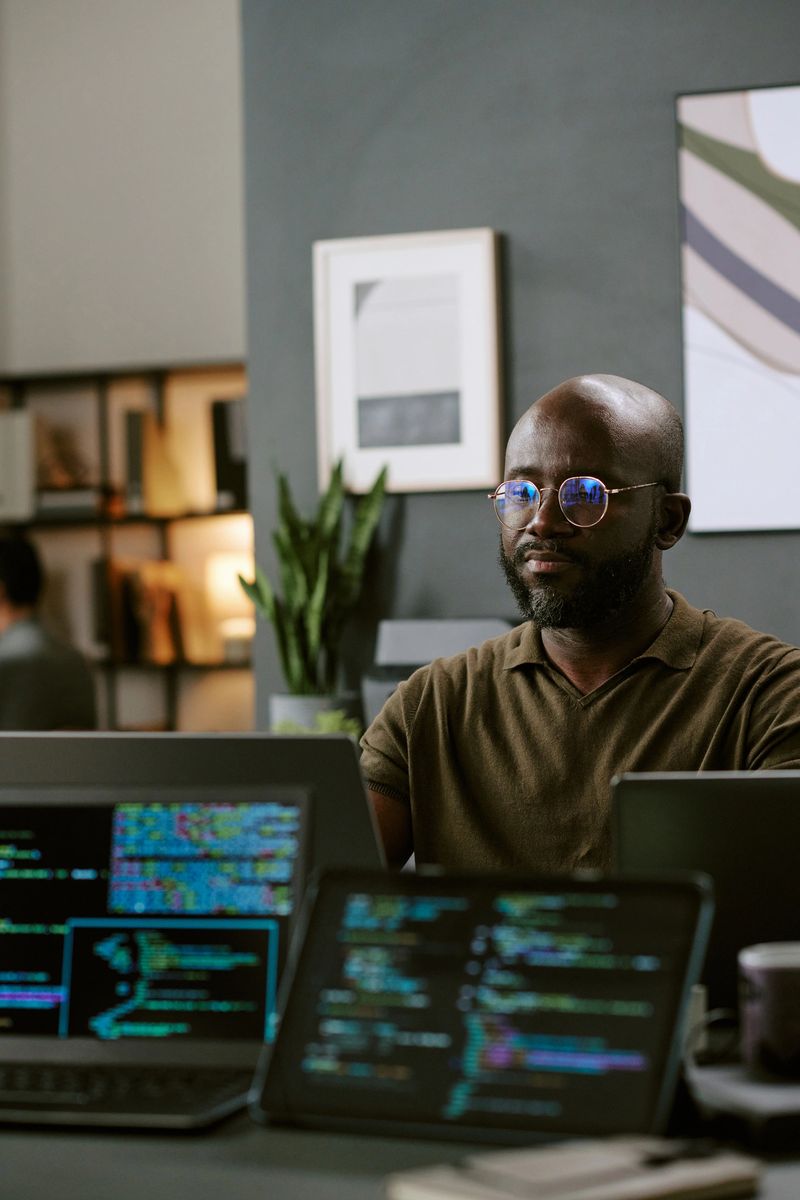Young Black man wearing glasses working on multiple computer screens displaying programming code in modern office setting, focusing intently on digital tasks