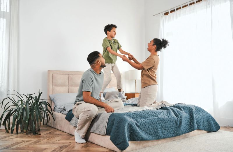 Family in a cozy home interior, sitting on a large comfortable bed in pajamas, holding their son by the hands while he jumps on the bed, enjoying a cozy and fun morning together in a bright bedroom.