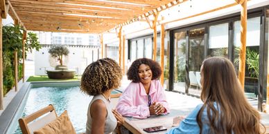 Three women chatting happily by a poolside patio under string lights.