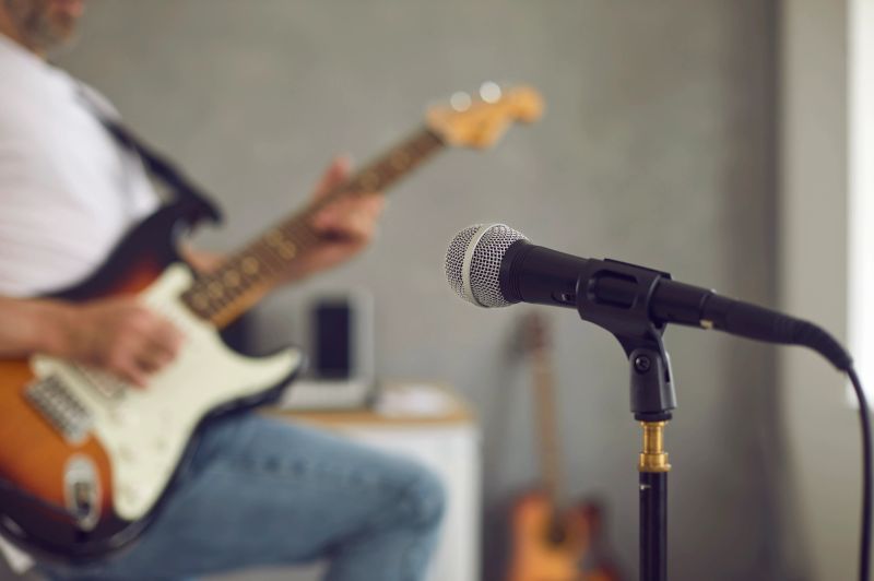 Close up of microphone in recording studio or rehearsal room, with man playing some loud music on electric or bass guitar in blurred background. Modern mic shot in soft, selective focus. Music concept