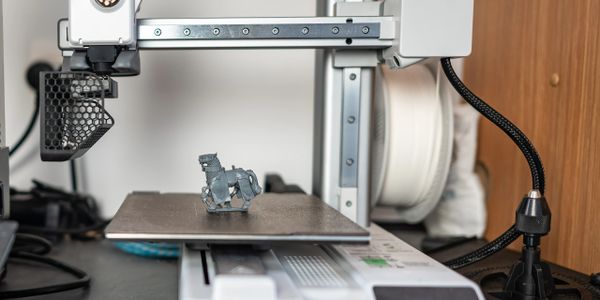 3D printer creating a small grey horse figurine on a printing bed.