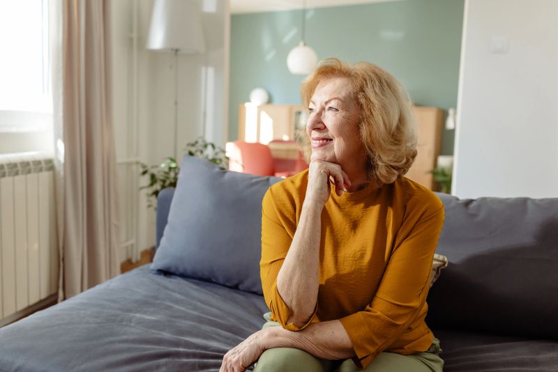 A senior woman sits on a comfortable sofa in a modern living room, resting her chin on her hand with a thoughtful expression. Warm light highlights a cozy, relaxed home mood.