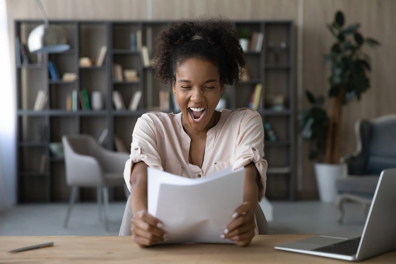 Happy excited African student girl receiving admission letter from university, college, holding and reading paper document, smiling, laughing, shouting for joy, celebrating achieve