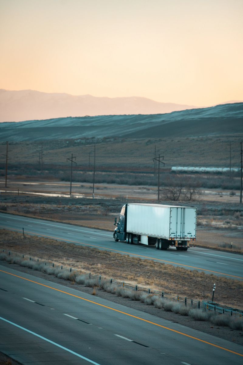 A white semi-truck with box trailer travels eastbound on a multi-lane highway through the open Utah desert. The truck is viewed from behind as it moves along the right lane, silhouetted against rolling brown hills and distant mountains bathed in soft orange-pink sunrise light. Power lines, sparse dry vegetation, and a faint train in the far background emphasize the vast, isolated Western landscape at dawn.