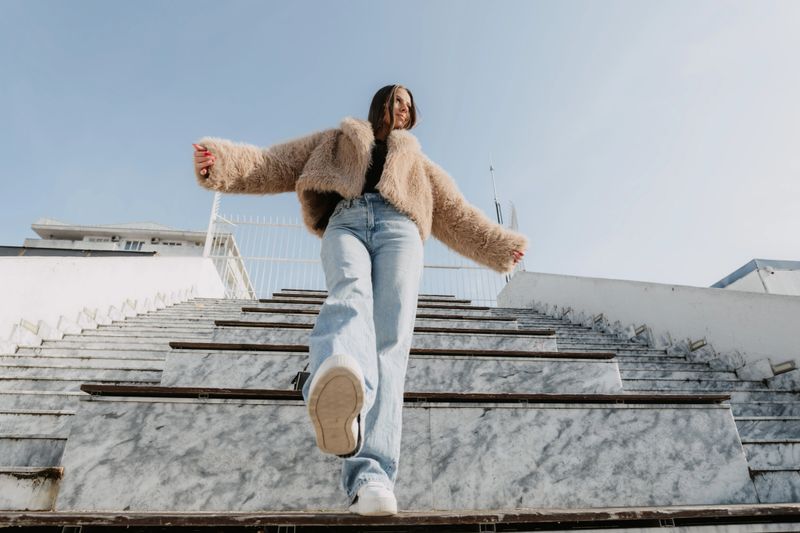 Playful and confident young woman posing on marble stairs in an urban environment, wearing a stylish winter coat and casual jeans. Fashion lifestyle concept with modern architecture, natural daylight and expressive body language.