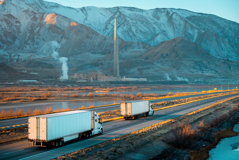 Two white semi-trucks, towing a white trailer, zoom down a multi-lane highway in the stark Utah landscape. Behind them, a large industrial complex—probably a power plant or refinery—stands tall, its smokestacks belching thick white steam or vapor into the cold, overcast sky. Snow-dusted mountains rise in the distance, framing the scene with barren winter brush and wetlands along the roadside. The mood is a mix of industrial grit and rugged Western expanse, all captured in cool, muted tones.