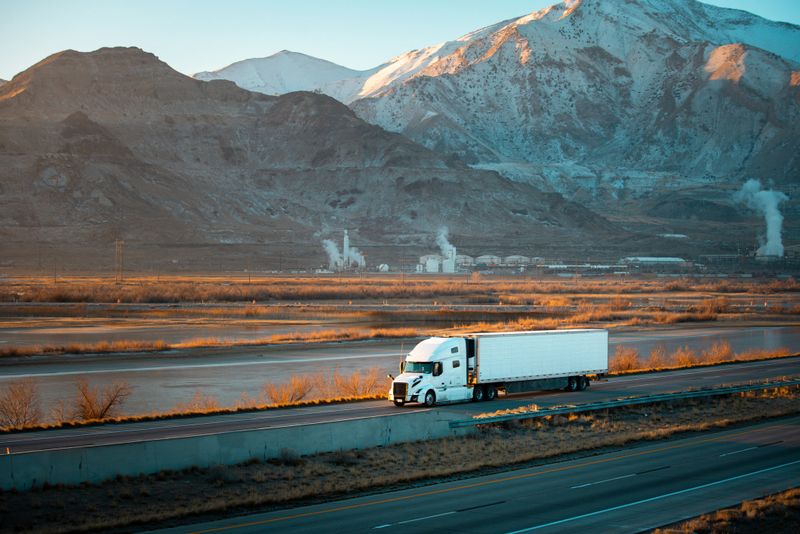 A white semi-truck with a white trailer speeds along a multi-lane highway in the stark Utah landscape. Behind it, a large industrial complex—likely a power plant or refinery—rises, its tall smokestacks emitting thick white steam or vapor into the cold, overcast sky. Snow-dusted mountains loom in the distance, framing the scene with barren winter brush and wetlands along the roadside. The mood is a blend of industrial grit and rugged Western expanse, captured in cool, muted tones.