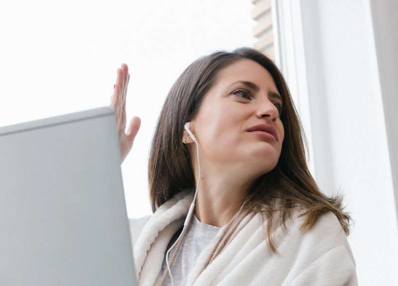 Woman working from home in a bathrobe, using a laptop with earphones during a virtual call. Ideal for remote work, online communication, home office lifestyle, and flexible schedule concepts.