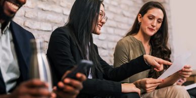 Two women reviewing a document with a man using a phone nearby.