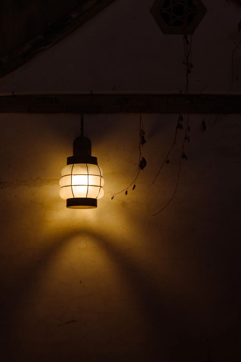 A classic kerosene lantern hangs on a wall, its warm, dim yellow light casting a soft, atmospheric glow and gentle shadows on the surrounding surface.