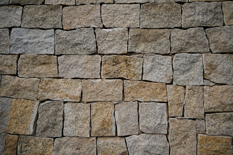 A close-up view of a hand-built stone wall made from natural granite blocks in varying shades of gray, beige, and brown. The stones are irregularly shaped and carefully fitted together, creating a rustic and durable surface. This image captures the natural texture, grain, and weathered appearance of traditional masonry work, ideal for use as a background, texture overlay, or architectural reference