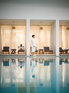 People relaxing by a poolside seating area with curtains and reflections in the water.