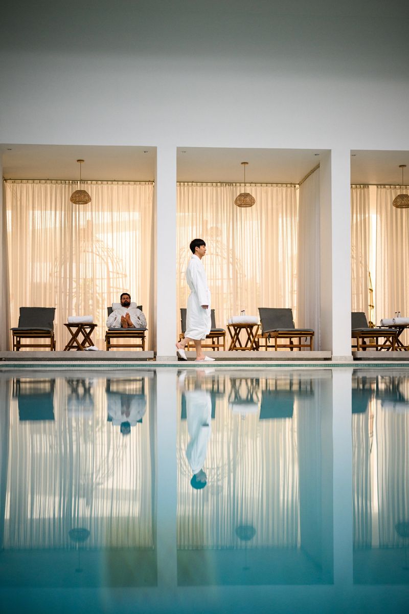 Full length shot of an elegant indoor hotel pool with symmetrical spa cabanas, lounge chairs and sheer golden curtains, with still water reflections and a luxury wellness atmosphere.