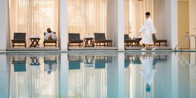 Person relaxing by an indoor pool with another walking by.