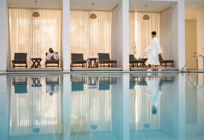 Wide shot of a hotel spa interior showing a man in a white robe walking beside the pool, with lounge chairs, sheer golden curtains and mirrored water reflections.