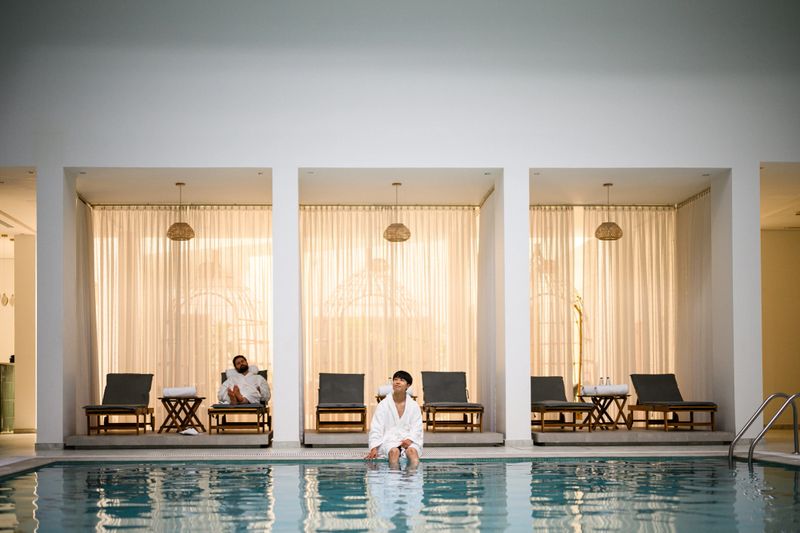Wide shot of a luxury indoor hotel pool showing a man in a white robe dipping legs in shallow water, with private spa cabanas, golden curtains and modern architecture behind him.