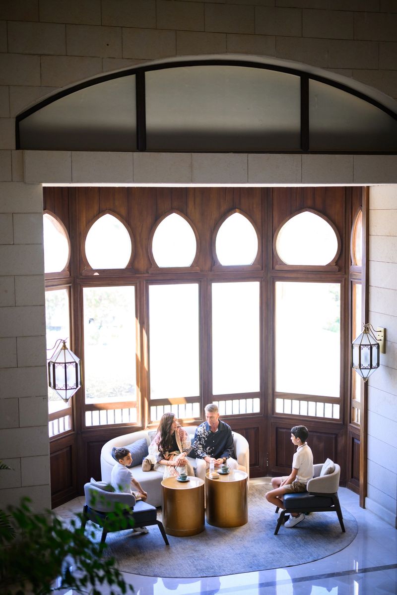 High angle view of a luxury hotel lobby lounge showing a family sitting on a couch and armchairs, with arched wooden windows, stone walls and sunlight in the background.