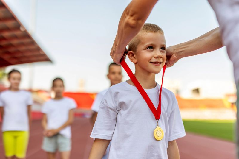 A smiling young boy in a white T-shirt wears a gold medal on a red ribbon after a track event. Team members cheer in the background, celebrating achievement and teamwork.