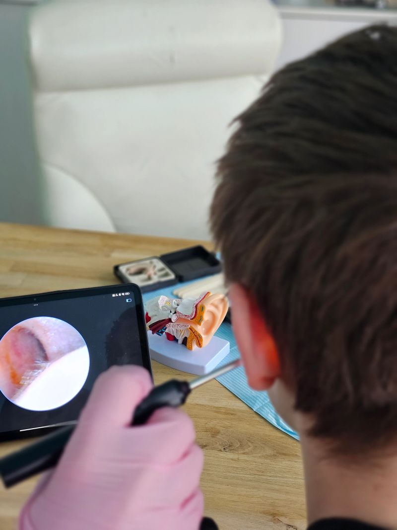 A young person examines an ear on a tablet screen while holding an otoscope. A model of an ear is on the table, indicating a medical or educational setting.