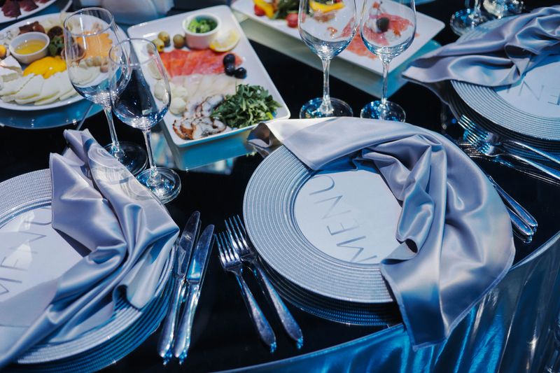 Elegant table setting prepared for a formal event or banquet. Close-up of neatly arranged plates with menus, silver cutlery, folded satin napkins, and empty wine glasses. Luxury catering concept.