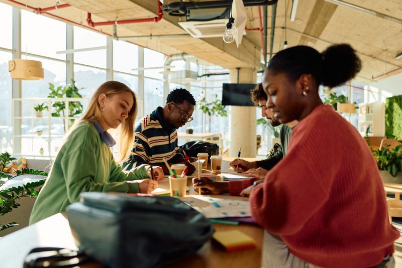 Group of young adult students collaborating on a project, writing notes and discussing ideas at a brightly lit desk in a contemporary university coworking space