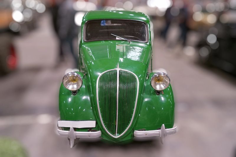 A classic green car stands on display at a vintage vehicle exhibition in a large indoor space. Visitors walk around to admire the cars at the event.