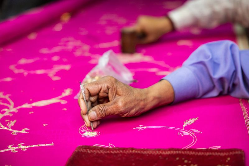Close-up of an artisan's weathered hand carefully applying gold thread embroidery onto vibrant magenta fabric. Using a small tool, the craftsperson creates intricate floral and paisley motifs with precise, delicate stitching that showcases traditional handwork and textile artistry.