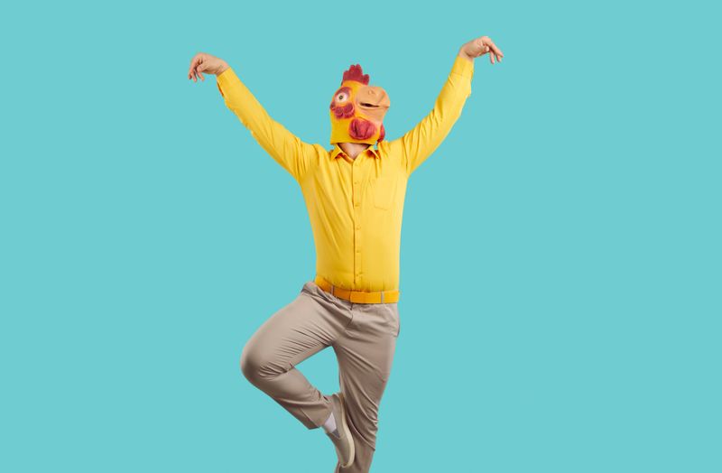 Cheerful young man in rubber rooster mask shows funny dance moves on turquoise background. Man with a chicken mask on his head, wearing yellow shirt and pants having fun on colored background.