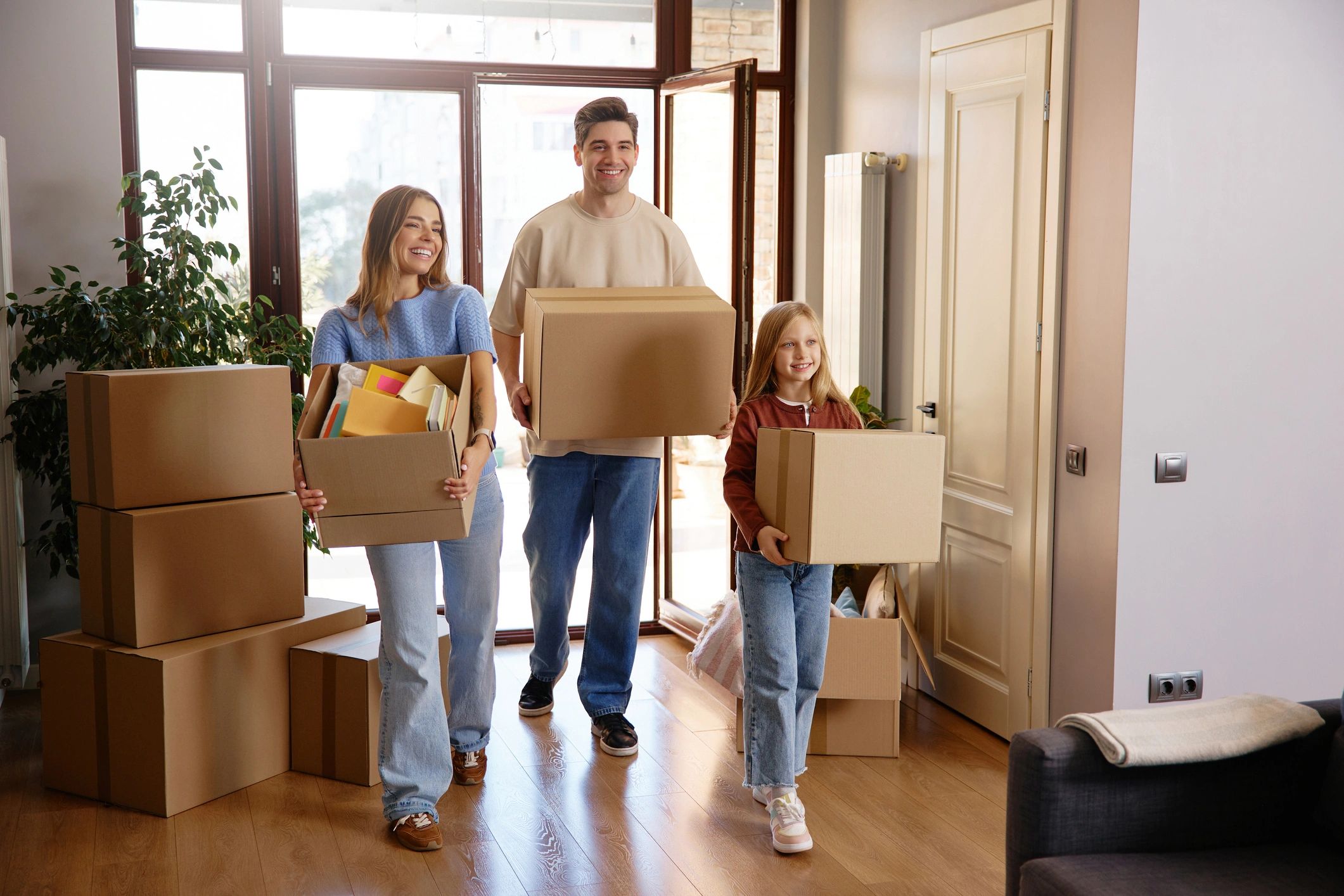 A happy family of three carrying cardboard boxes into a bright, modern new home. A smiling man, woma