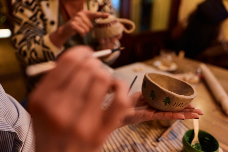 Woman hands holding a small, unglazed ceramic bowl decorated with painted green pine trees, focusing intently on the artistic craft during a pottery class