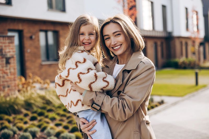 Happy mother holding her young daughter while standing and smiling together outdoors in residential neighborhood, representing family bonding and joyful lifestyle. Love, childhood concept