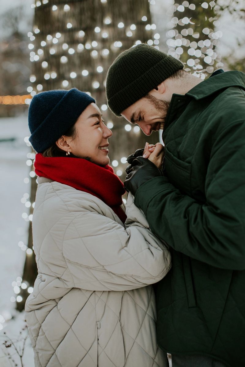 A Korean woman and a Caucasian man smile at each other while standing close together. They are dressed warmly and surrounded by trees with lights. It is winter and they appear happy.