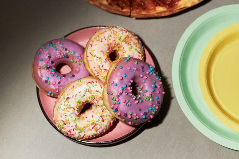 Plate holding four donuts with pink and purple icing topped with colorful sprinkles, sitting on table near pastel green and yellow paper plate and partially visible pizza