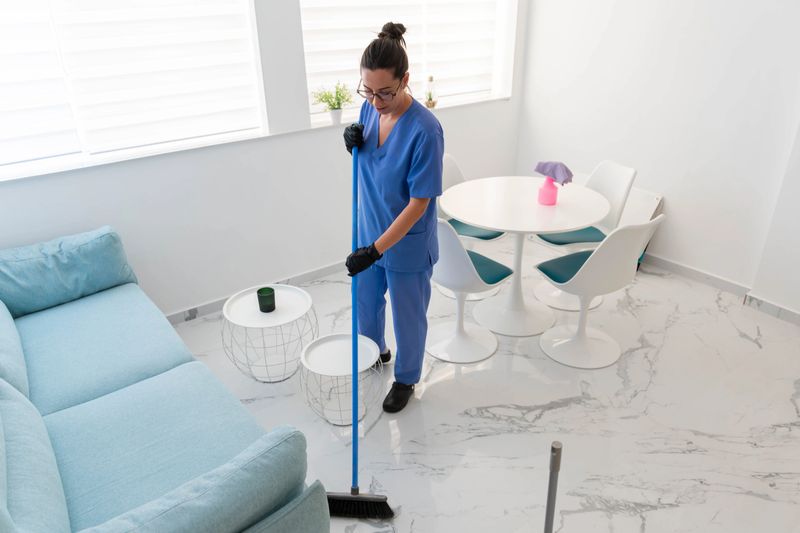 Housecleaner in blue uniform and black gloves meticulously wipes a pristine white marble floor in a bright modern apartment, ensuring thorough hygiene and tidiness
