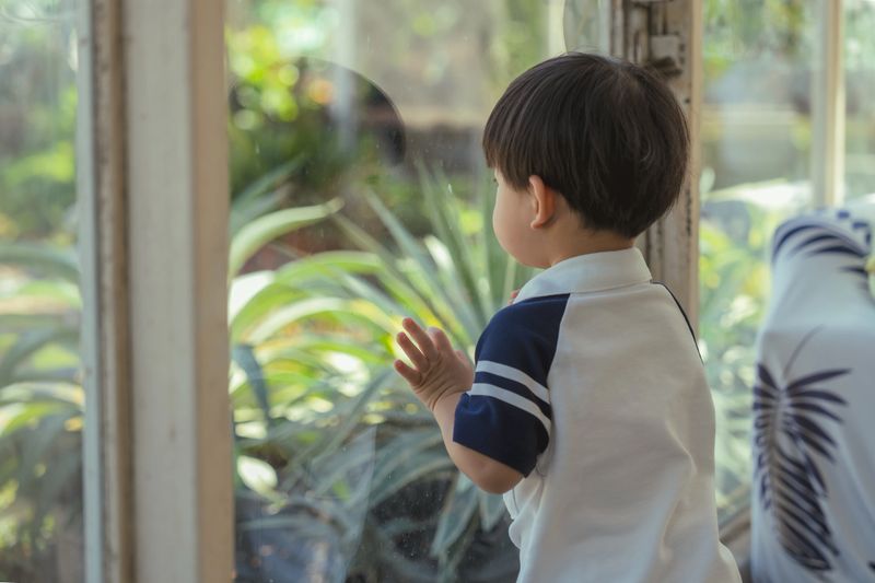 curious Asian child standing peacefully by the large glass window looking out at the beautiful green garden plants outside, placing his small hand gently on the glass with wonder and contemplation, enjoying the serene natural view