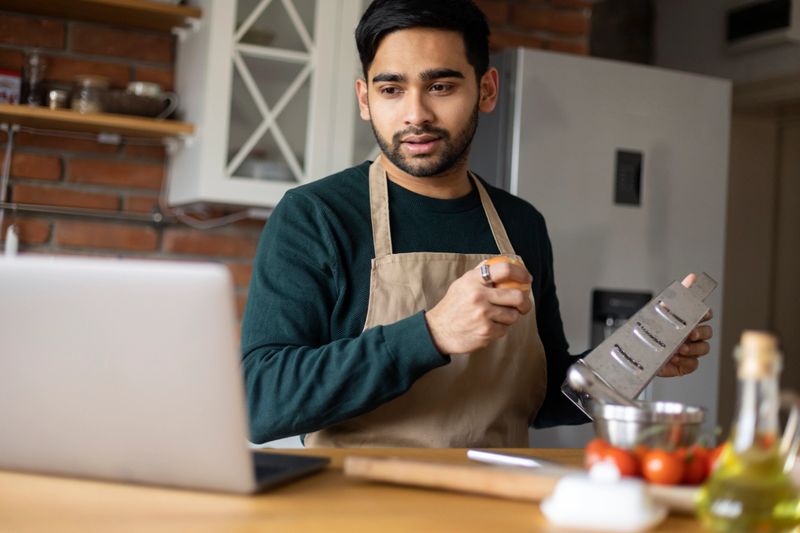 Young man preparing food in a kitchen using a laptop. About 25 years old, Indian male.