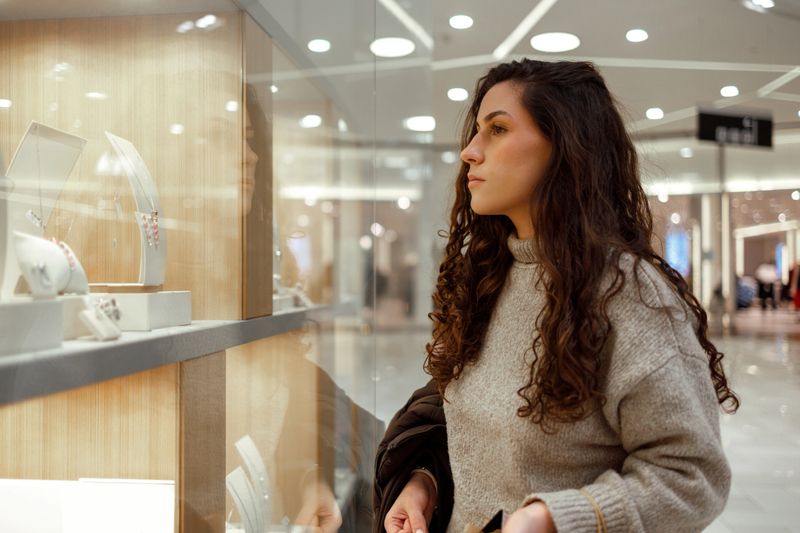 Young woman standing in a shopping mall and looking at jewelry displayed in a store window inside a retail center.