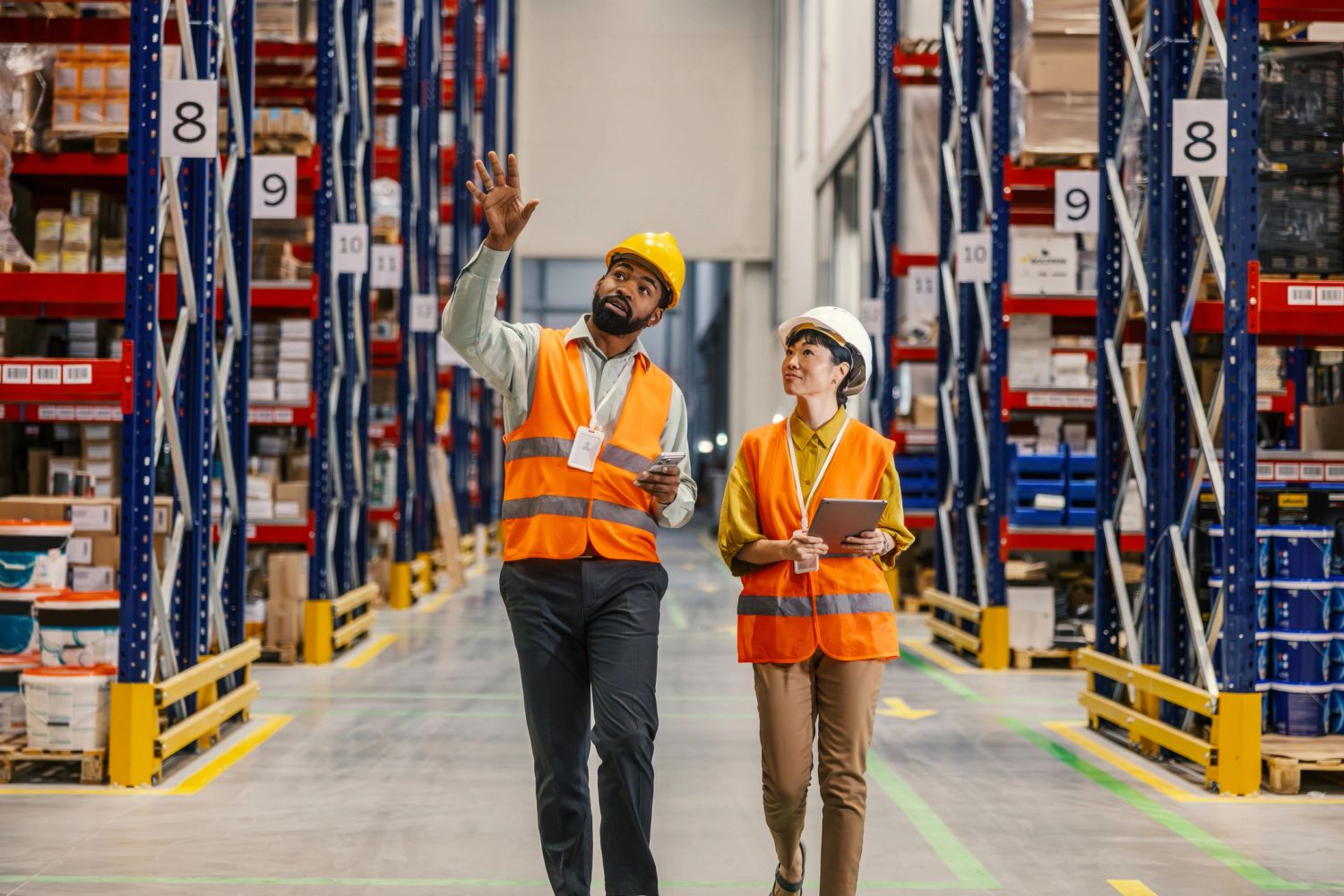 Two warehouse workers discuss stock while walking through aisles.