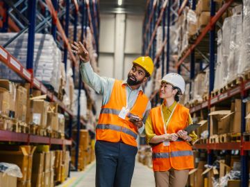 Two warehouse workers in safety gear discussing inventory.