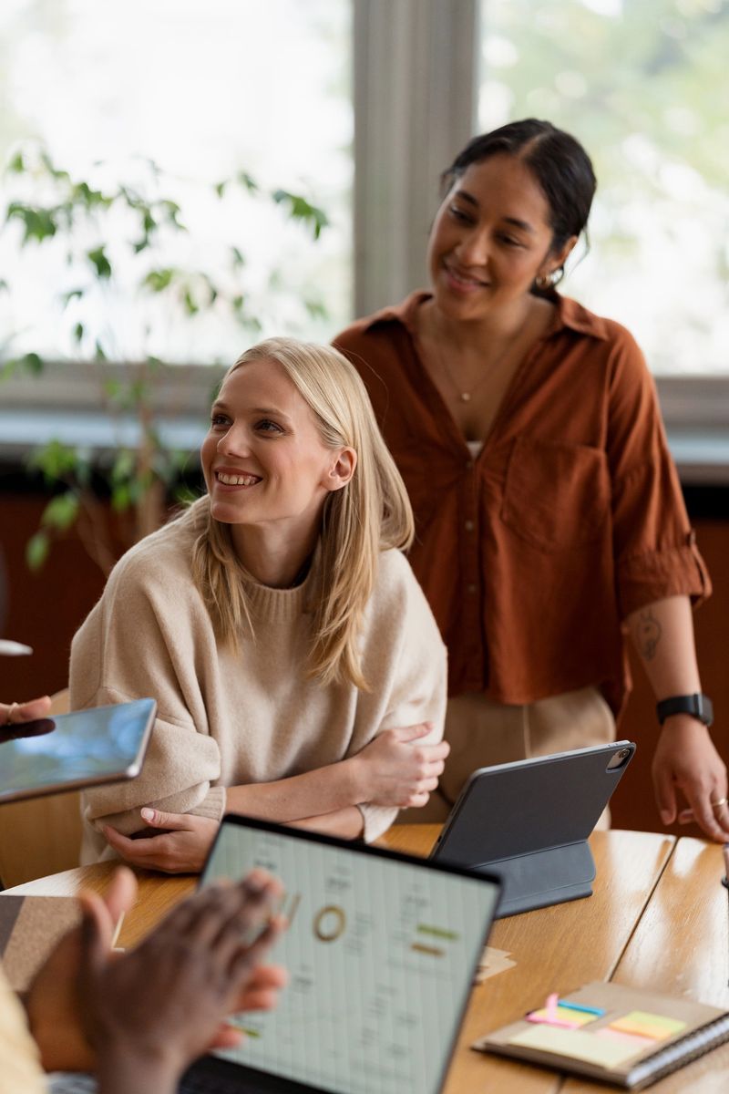 Diverse businesswomen smiling, discussing ideas and collaborating during a meeting in a modern office setup, using screens and devices