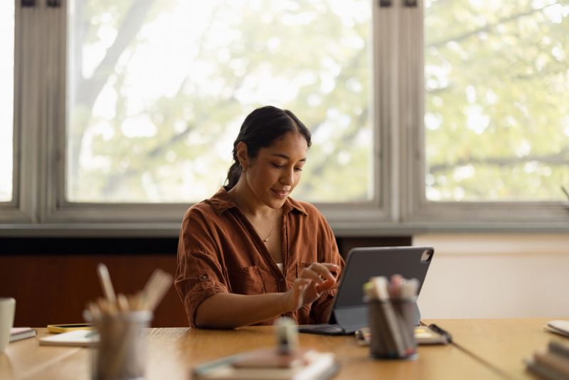 Young adult woman concentrating and smiling while working or studying indoors using a digital tablet at a wooden desk