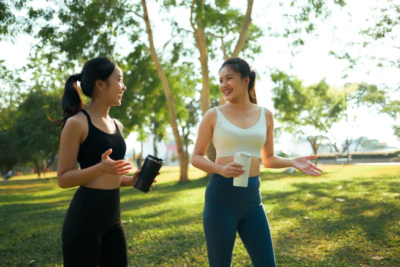 Two smiling young adult women in activewear standing on a sunny park lawn, holding water bottles and talking after a workout
