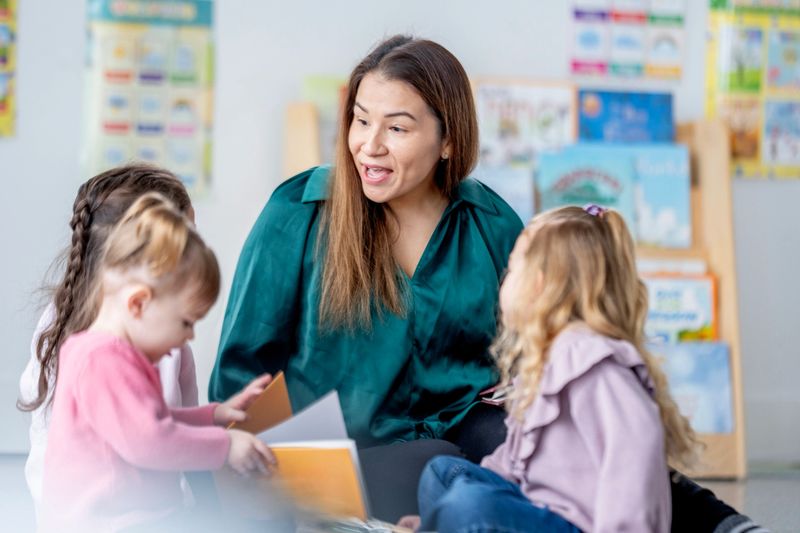 A caring teacher guides a group of young children as they explore colorful books in a bright classroom. The scene conveys learning, collaboration, and a warm educational environment.
